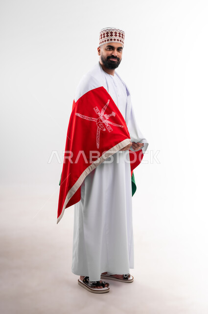 Participation in national holidays and occasions, celebration of Omani National Day, pride in Omani symbols and flag, portrait of a young Gulf Arab Omani man wearing a dishdasha and a kuma, concept of love of the homeland and belonging to it, full body, white background