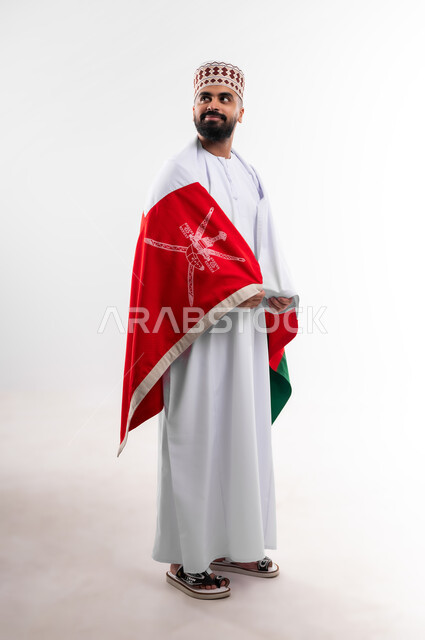 Participation in national holidays and occasions, celebration of Omani National Day, pride in Omani symbols and flag, portrait of a young Gulf Arab Omani man wearing a dishdasha and a kuma, concept of love of the homeland and belonging to it, full body, white background