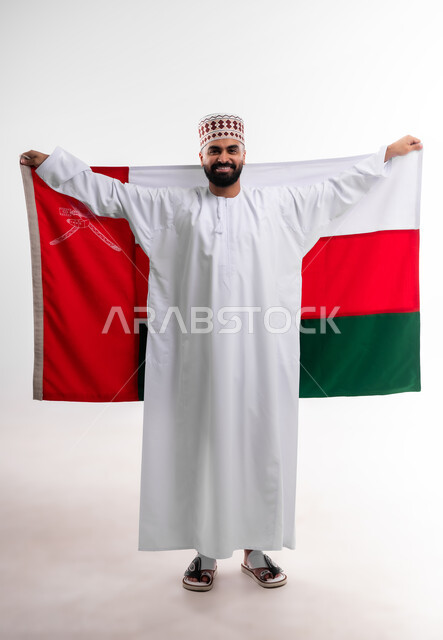 Participation in national holidays and occasions, celebration of Omani National Day, pride in Omani symbols and flag, portrait of a young Gulf Arab Omani man wearing a dishdasha and a kuma, concept of love of the homeland and belonging to it, full body, white background