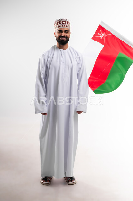 المشاركة بالاعياد والمناسبات الوطنية، الاحتفال بذكرى اليوم الوطني العماني، الاعتزاز والفخر بالرموز والراية العمانية، بورتريه لشاب عربي خليجي عماني يرتدي الدشداشة والكمه، مفهوم حب الوطن والإنتماء إليه، الجسم بالطول الكامل، خلفية بيضاء