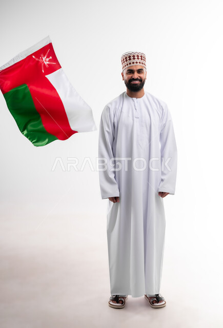 المشاركة بالاعياد والمناسبات الوطنية، الاحتفال بذكرى اليوم الوطني العماني، الاعتزاز والفخر بالرموز والراية العمانية، بورتريه لشاب عربي خليجي عماني يرتدي الدشداشة والكمه، مفهوم حب الوطن والإنتماء إليه، الجسم بالطول الكامل، خلفية بيضاء