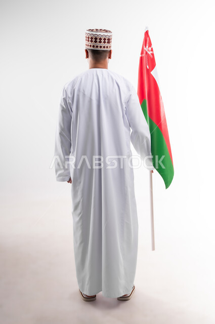 المشاركة بالاعياد والمناسبات الوطنية، الاحتفال بذكرى اليوم الوطني العماني، الاعتزاز والفخر بالرموز والراية العمانية، بورتريه لشاب عربي خليجي عماني يرتدي الدشداشة والكمه، مفهوم حب الوطن والإنتماء إليه، الجسم بالطول الكامل، خلفية بيضاء