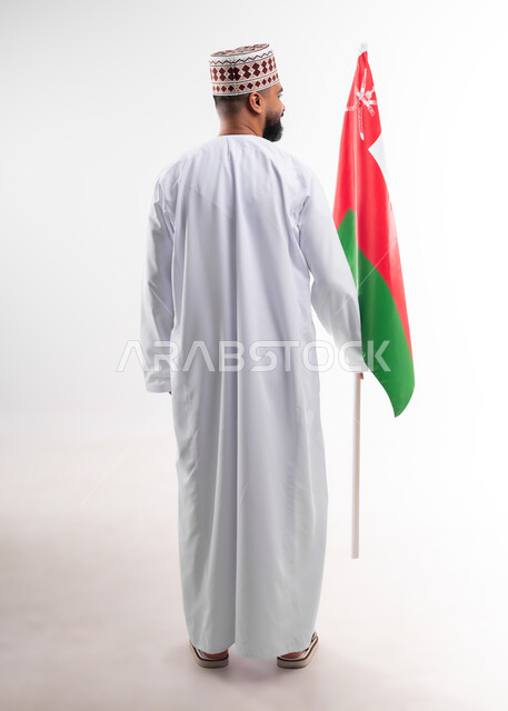 المشاركة بالاعياد والمناسبات الوطنية، الاحتفال بذكرى اليوم الوطني العماني، الاعتزاز والفخر بالرموز والراية العمانية، بورتريه لشاب عربي خليجي عماني يرتدي الدشداشة والكمه، مفهوم حب الوطن والإنتماء إليه، الجسم بالطول الكامل، خلفية بيضاء