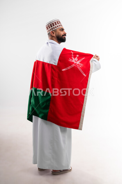 Participation in national holidays and occasions, celebration of Omani National Day, pride in Omani symbols and flag, portrait of a young Gulf Arab Omani man wearing a dishdasha and a kuma, concept of love of the homeland and belonging to it, full body, white background