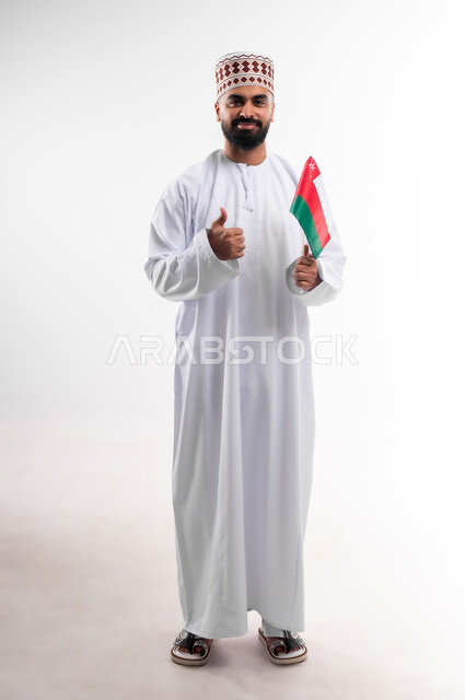 المشاركة بالاعياد والمناسبات الوطنية، الاحتفال بذكرى اليوم الوطني العماني، الاعتزاز والفخر بالرموز والراية العمانية، بورتريه لشاب عربي خليجي عماني يرتدي الدشداشة والكمه، مفهوم حب الوطن والإنتماء إليه، الجسم بالطول الكامل، خلفية بيضاء