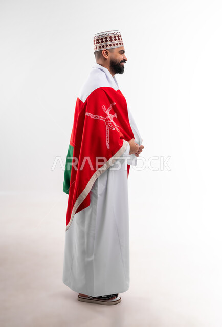 المشاركة بالاعياد والمناسبات الوطنية، الاحتفال بذكرى اليوم الوطني العماني، الاعتزاز والفخر بالرموز والراية العمانية، بورتريه لشاب عربي خليجي عماني يرتدي الدشداشة والكمه، مفهوم حب الوطن والإنتماء إليه، الجسم بالطول الكامل، خلفية بيضاء