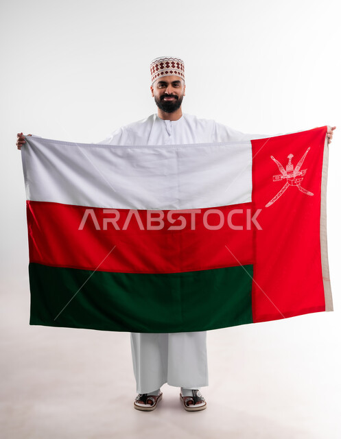 المشاركة بالاعياد والمناسبات الوطنية، الاحتفال بذكرى اليوم الوطني العماني، الاعتزاز والفخر بالرموز والراية العمانية، بورتريه لشاب عربي خليجي عماني يرتدي الدشداشة والكمه، مفهوم حب الوطن والإنتماء إليه، الجسم بالطول الكامل، خلفية بيضاء
