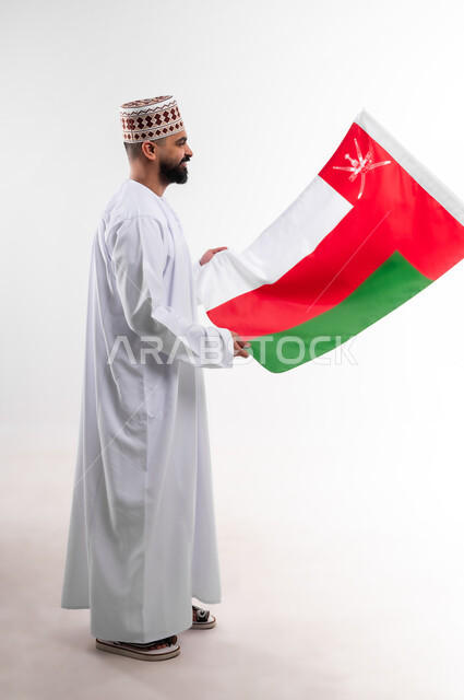 Participation in national holidays and occasions, celebration of Omani National Day, pride in Omani symbols and flag, portrait of a young Gulf Arab Omani man wearing a dishdasha and a kuma, concept of love of the homeland and belonging to it, full body, white background