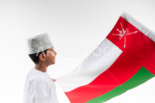 الفخر والاعتزاز بالانتماء للهوية، المشاركة بالاعياد والمناسبات الوطنية، الاحتفال بذكرى اليوم الوطني العماني، الاعتزاز والفخر بالرموز والراية العمانية، بورتريه لطفل عربي خليجي عماني يرتدي الدشداشة والكمه، مفهوم حب الوطن، خلفية بيضاء