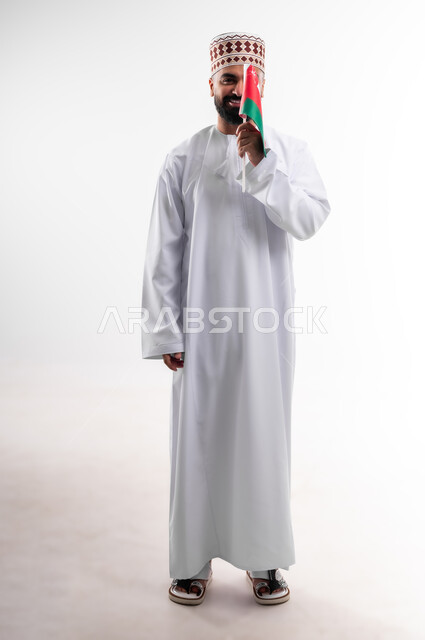 Participation in national holidays and occasions, celebration of Omani National Day, pride in Omani symbols and flag, portrait of a young Gulf Arab Omani man wearing a dishdasha and a kuma, concept of love of the homeland and belonging to it, full body, white background