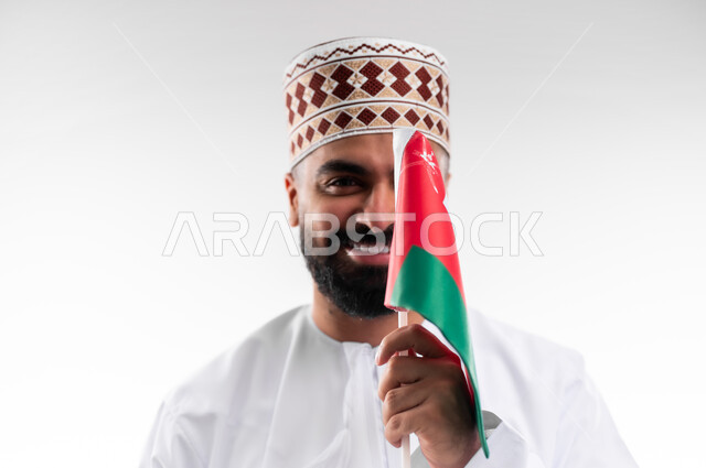 الفخر والاعتزاز بالانتماء للهوية، المشاركة بالاعياد والمناسبات الوطنية، الاحتفال بذكرى اليوم الوطني العماني، الاعتزاز والفخر بالرموز والراية العمانية، بورتريه لشاب عربي خليجي عماني يرتدي الدشداشة والكمه، مفهوم حب الوطن، خلفية بيضاء
