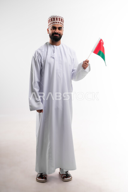 Participation in national holidays and occasions, celebration of Omani National Day, pride in Omani symbols and flag, portrait of a young Gulf Arab Omani man wearing a dishdasha and a kuma, concept of love of the homeland and belonging to it, full body, white background