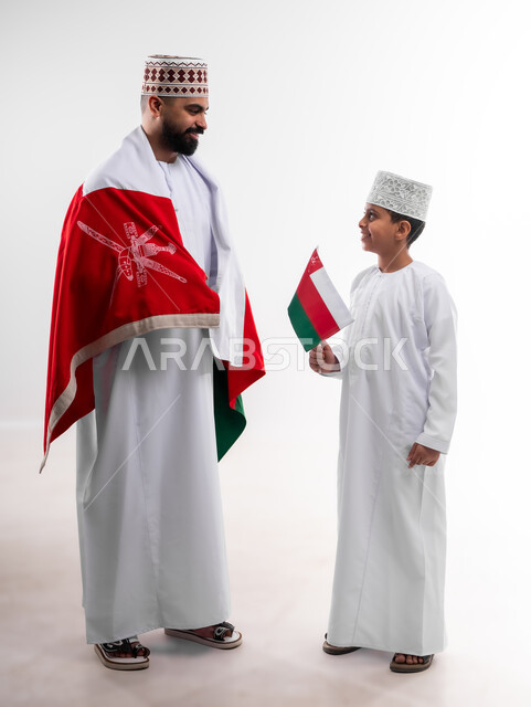 الاحتفال بذكرى اليوم الوطني العماني، الرايات والأعلام الوطنية للدول العربية، الاعتزاز والفخر بالرموز والراية العمانية، بورتريه لطفل وشاب عربي خليجي عماني يرتدي الدشداشة والكمه، مفهوم حب الوطن والإنتماء إليه، الجسم بالطول الكامل، خلفية بيضاء