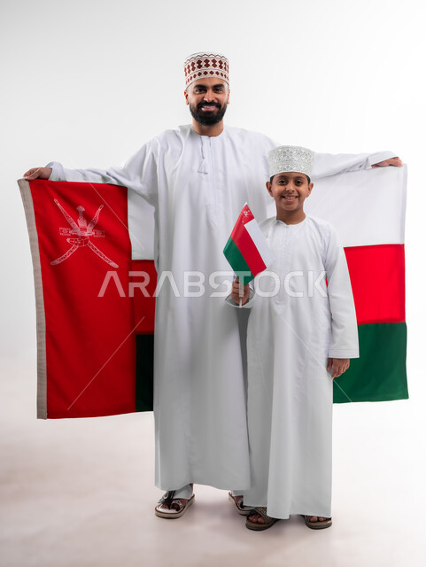 الاحتفال بذكرى اليوم الوطني العماني، الرايات والأعلام الوطنية للدول العربية، الاعتزاز والفخر بالرموز والراية العمانية، بورتريه لطفل وشاب عربي خليجي عماني يرتدي الدشداشة والكمه، مفهوم حب الوطن والإنتماء إليه، الجسم بالطول الكامل، خلفية بيضاء