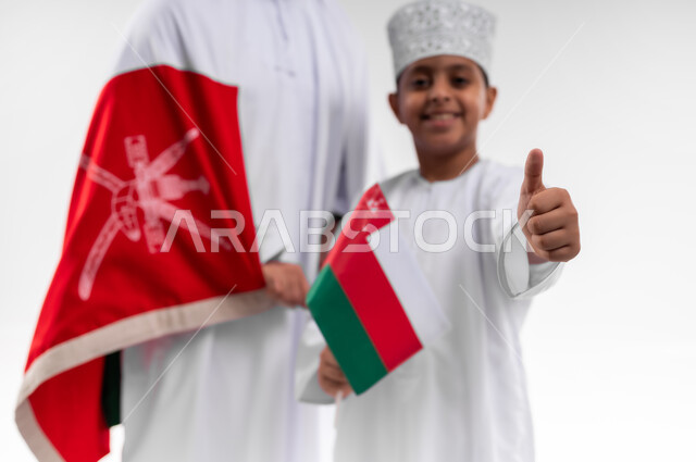 الاحتفال بذكرى اليوم الوطني العماني، الرايات والأعلام الوطنية للدول العربية، الاعتزاز والفخر بالرموز والراية العمانية، بورتريه مقرب لطفل وشاب عربي خليجي عماني يرتدي الدشداشة والكمه، مفهوم حب الوطن والإنتماء إليه، خلفية بيضاء
