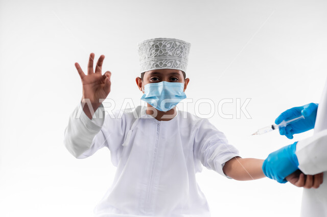 اتباع قوانين وزارة الصحة، اتاحة تلقي التطعيم ضد فايروس كورونا للاطفال، بورتريه مقرب لطبيب يرتدي المعطف والقفازات الطبية، يقوم باعطاء جرعة لقاح كوفيد - 19 لطفل عربي خليجي عماني، الالتزام بإجراءات السلامة، خلفية بيضاء