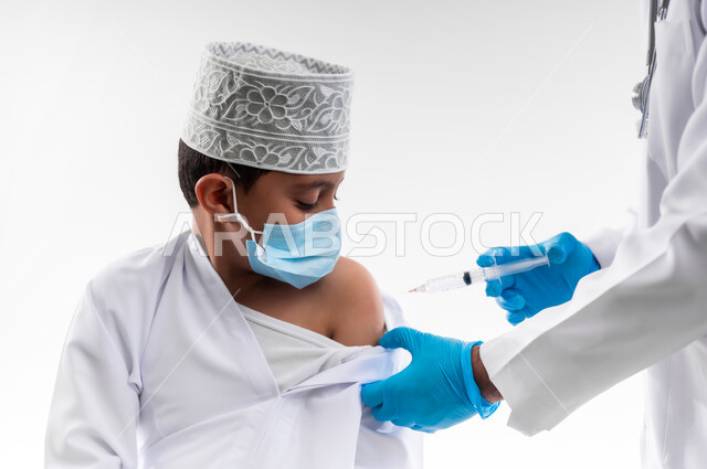 اتباع قوانين وزارة الصحة، اتاحة تلقي التطعيم ضد فايروس كورونا للاطفال، بورتريه مقرب لطبيب يرتدي المعطف والقفازات الطبية، يقوم باعطاء جرعة لقاح كوفيد - 19 لطفل عربي خليجي عماني، الالتزام بإجراءات السلامة، خلفية بيضاء