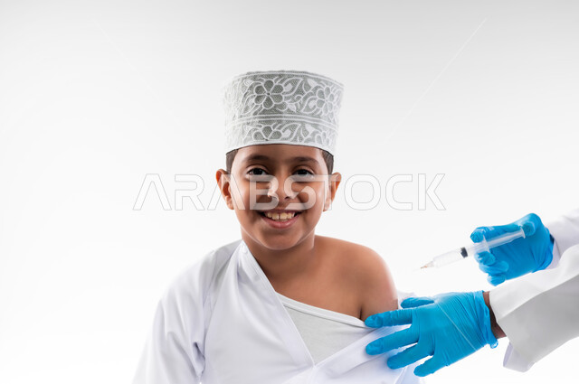 اتباع قوانين وزارة الصحة، اتاحة تلقي التطعيم ضد فايروس كورونا للاطفال، بورتريه مقرب لطبيب يرتدي المعطف والقفازات الطبية، يقوم باعطاء جرعة لقاح كوفيد - 19 لطفل عربي خليجي عماني، الالتزام بإجراءات السلامة، الجسم بالطول الكامل، خلفية بيضاء