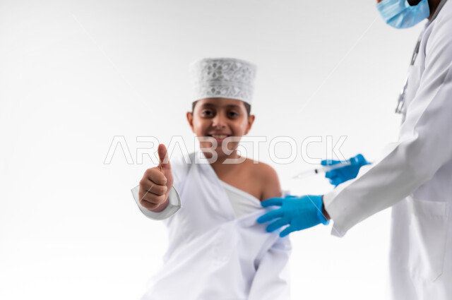 اتباع قوانين وزارة الصحة، اتاحة تلقي التطعيم ضد فايروس كورونا للاطفال، بورتريه مقرب لطبيب يرتدي المعطف والقفازات الطبية، يقوم باعطاء جرعة لقاح كوفيد - 19 لطفل عربي خليجي عماني، الالتزام بإجراءات السلامة، الجسم بالطول الكامل، خلفية بيضاء