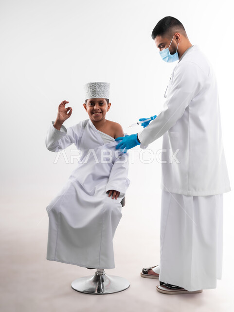 اتاحة تلقي التطعيم ضد فايروس كورونا للاطفال، بورتريه لطبيب يرتدي المعطف والقفازات الطبية ، يقوم باعطاء جرعة لقاح كوفيد - 19 لطفل عربي خليجي عماني ، الالتزام بإجراءات السلامة ، اتباع قوانين وزارة الصحة ، الجسم بالطول الكامل، خلفية بيضاء