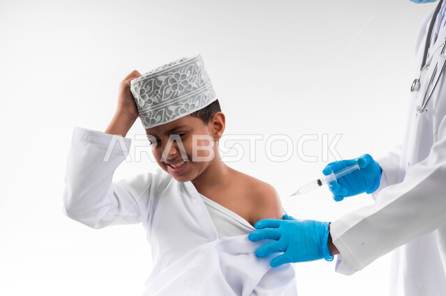 اتباع قوانين وزارة الصحة، اتاحة تلقي التطعيم ضد فايروس كورونا للاطفال، بورتريه مقرب لطبيب يرتدي المعطف والقفازات الطبية، يقوم باعطاء جرعة لقاح كوفيد - 19 لطفل عربي خليجي عماني، الالتزام بإجراءات السلامة، الجسم بالطول الكامل، خلفية بيضاء