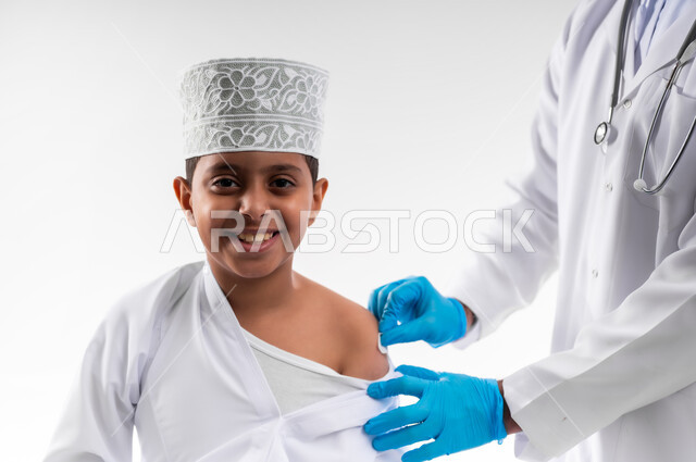 اتباع قوانين وزارة الصحة، اتاحة تلقي التطعيم ضد فايروس كورونا للاطفال، بورتريه مقرب لطبيب يرتدي المعطف والقفازات الطبية، يقوم باعطاء جرعة لقاح كوفيد - 19 لطفل عربي خليجي عماني، الالتزام بإجراءات السلامة، الجسم بالطول الكامل، خلفية بيضاء