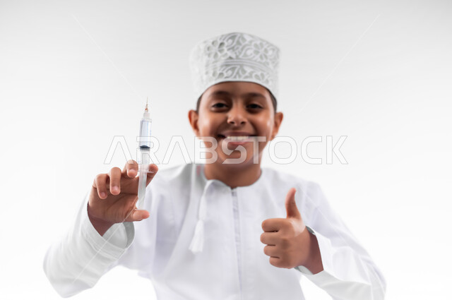 مفهوم الطب و الرعاية الصحية الأولية، تحديد مهنة المستقبل منذ الصغر، طموحات الأطفال في العمل بالمجال الطبي، بورتريه مقرب لطفل عربي خليجي عماني مبتسم يرتدي الدشداشة والكمه  ويحمل بيده إبرة،  مهنة إنسانية واجتماعية، خلفية بيضاء
