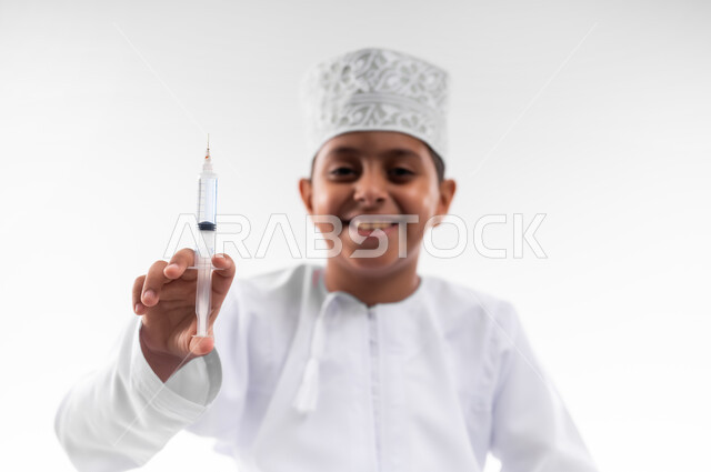 مفهوم الطب و الرعاية الصحية الأولية، تحديد مهنة المستقبل منذ الصغر، طموحات الأطفال في العمل بالمجال الطبي، بورتريه مقرب لطفل عربي خليجي عماني مبتسم يرتدي الدشداشة والكمه  ويحمل بيده إبرة،  مهنة إنسانية واجتماعية، خلفية بيضاء