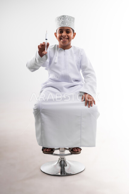 تحديد مهنة المستقبل منذ الصغر، طموحات الأطفال في العمل بالمجال الطبي، بورتريه لطفل عربي خليجي عماني مبتسم يرتدي الدشداشة والكمه  ويحمل بيده إبرة، مفهوم الطب و الرعاية الصحية الأولية ، مهنة إنسانية واجتماعية، الجسم بالطول الكامل، خلفية بيضاء