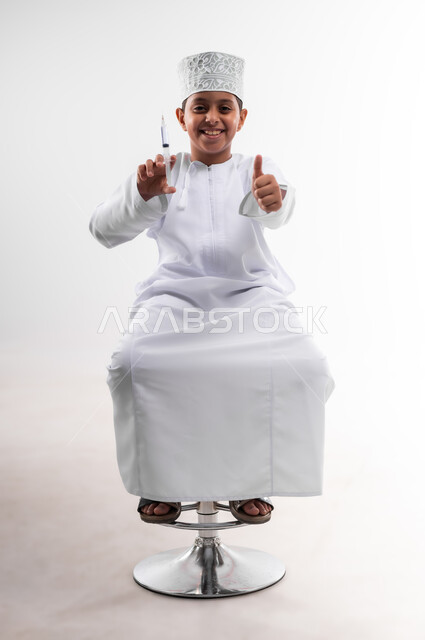 تحديد مهنة المستقبل منذ الصغر، طموحات الأطفال في العمل بالمجال الطبي، بورتريه لطفل عربي خليجي عماني مبتسم يرتدي الدشداشة والكمه  ويحمل بيده إبرة، مفهوم الطب و الرعاية الصحية الأولية ، مهنة إنسانية واجتماعية، الجسم بالطول الكامل، خلفية بيضاء