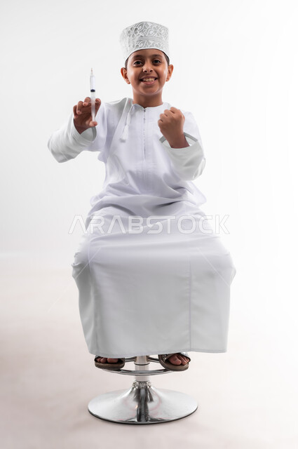 تحديد مهنة المستقبل منذ الصغر، طموحات الأطفال في العمل بالمجال الطبي، بورتريه لطفل عربي خليجي عماني مبتسم يرتدي الدشداشة والكمه  ويحمل بيده إبرة، مفهوم الطب و الرعاية الصحية الأولية ، مهنة إنسانية واجتماعية، الجسم بالطول الكامل، خلفية بيضاء