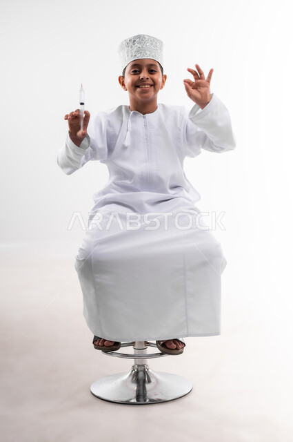 تحديد مهنة المستقبل منذ الصغر، طموحات الأطفال في العمل بالمجال الطبي، بورتريه لطفل عربي خليجي عماني مبتسم يرتدي الدشداشة والكمه  ويحمل بيده إبرة، مفهوم الطب و الرعاية الصحية الأولية ، مهنة إنسانية واجتماعية، الجسم بالطول الكامل، خلفية بيضاء