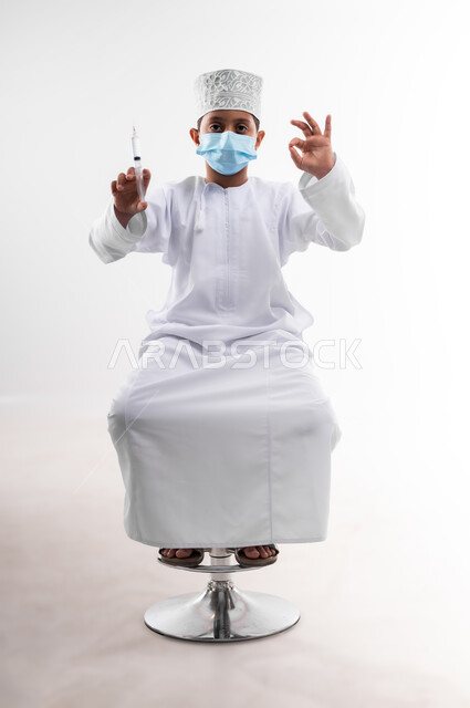 تحديد مهنة المستقبل منذ الصغر، طموحات الأطفال في العمل بالمجال الطبي، بورتريه لطفل عربي خليجي عماني مبتسم يرتدي الدشداشة   ويحمل بيده إبرة، مفهوم الطب و الرعاية الصحية الأولية، اتخاذ الإجراءات الاحترازية للوقاية من فايروس كورونا، خلفية بيضاء