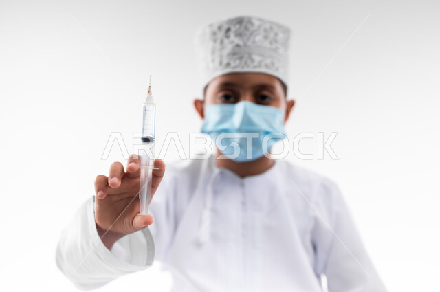 تحديد مهنة المستقبل منذ الصغر، بورتريه لطفل عربي خليجي عماني مبتسم يرتدي الكمامة والدشداشة والكمه ويحمل بيده إبرة، مفهوم الطب و الرعاية الصحية الأولية ، مهنة إنسانية واجتماعية، الإلتزام بالاجراءات الوقائية لفايروس كورونا، خلفية بيضاء