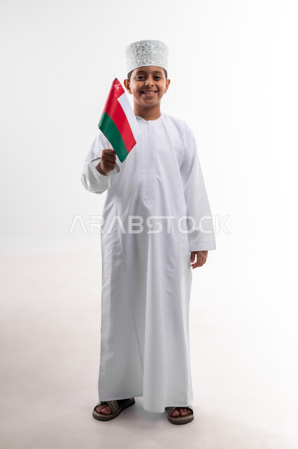 أعياد ومناسبات وطنية، إحياء ذكرى اليوم الوطني العماني، الاعتزاز والفخر بالرموز والراية العمانية، بورتريه لطفل عربي خليجي عماني يرتدي الدشداشة والكمه يحمل بيده علم صغير لسلطنة عمان، مفهوم حب الوطن والإنتماء إليه، الجسم بالطول الكامل، خلفية بيضاء