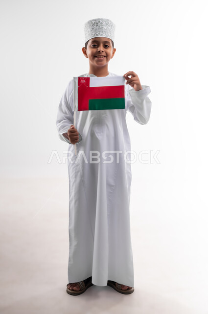 أعياد ومناسبات وطنية، إحياء ذكرى اليوم الوطني العماني، الاعتزاز والفخر بالرموز والراية العمانية، بورتريه لطفل عربي خليجي عماني يرتدي الدشداشة والكمه يحمل بيده علم صغير لسلطنة عمان، مفهوم حب الوطن والإنتماء إليه، الجسم بالطول الكامل، خلفية بيضاء