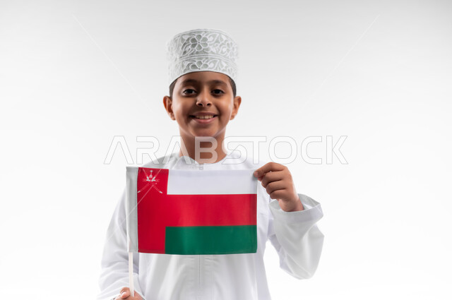 الرايات والأعلام الوطنية للدول العربية، إحياء ذكرى اليوم الوطني العماني، الاعتزاز والفخر بالرموز والراية العمانية، بورتريه مقرب لطفل عربي خليجي عماني يرتدي الدشداشة والكمه يحمل بيده علم صغير لسلطنة عمان، مفهوم حب الوطن والإنتماء إليه، خلفية بيضاء
