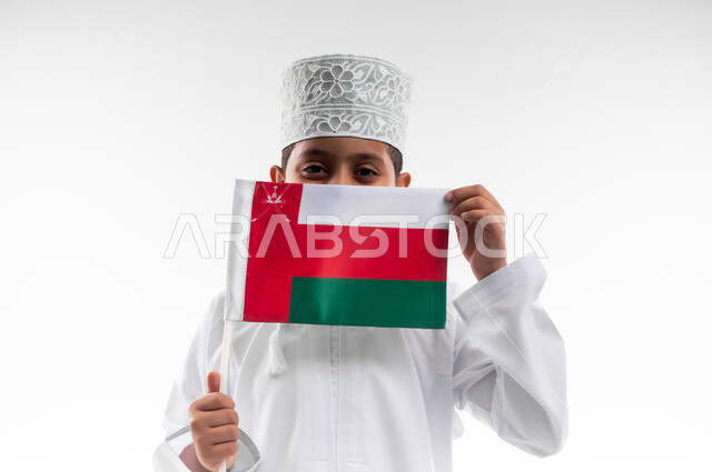 الرايات والأعلام الوطنية للدول العربية، إحياء ذكرى اليوم الوطني العماني، الاعتزاز والفخر بالرموز والراية العمانية، بورتريه مقرب لطفل عربي خليجي عماني يرتدي الدشداشة والكمه يحمل بيده علم صغير لسلطنة عمان، مفهوم حب الوطن والإنتماء إليه، خلفية بيضاء
