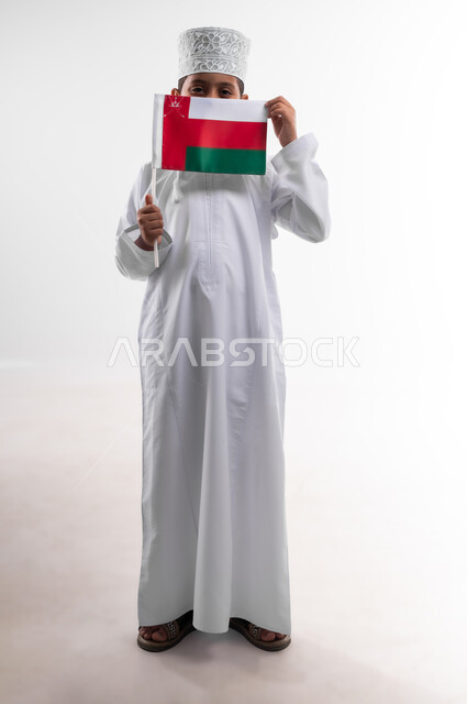 أعياد ومناسبات وطنية، إحياء ذكرى اليوم الوطني العماني، الاعتزاز والفخر بالرموز والراية العمانية، بورتريه لطفل عربي خليجي عماني يرتدي الدشداشة والكمه يحمل بيده علم صغير لسلطنة عمان، مفهوم حب الوطن والإنتماء إليه، الجسم بالطول الكامل، خلفية بيضاء
