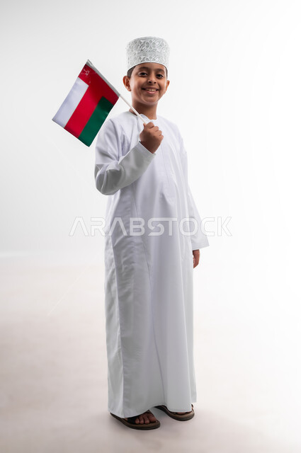 أعياد ومناسبات وطنية، إحياء ذكرى اليوم الوطني العماني، الاعتزاز والفخر بالرموز والراية العمانية، بورتريه لطفل عربي خليجي عماني يرتدي الدشداشة والكمه يحمل بيده علم صغير لسلطنة عمان، مفهوم حب الوطن والإنتماء إليه، الجسم بالطول الكامل، خلفية بيضاء