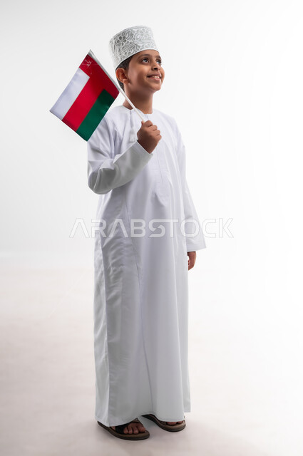 أعياد ومناسبات وطنية، إحياء ذكرى اليوم الوطني العماني، الاعتزاز والفخر بالرموز والراية العمانية، بورتريه لطفل عربي خليجي عماني يرتدي الدشداشة والكمه يحمل بيده علم صغير لسلطنة عمان، مفهوم حب الوطن والإنتماء إليه، الجسم بالطول الكامل، خلفية بيضاء