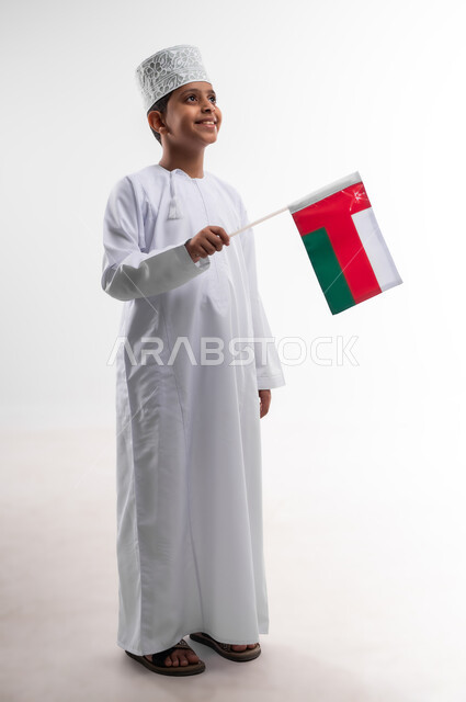 أعياد ومناسبات وطنية، إحياء ذكرى اليوم الوطني العماني، الاعتزاز والفخر بالرموز والراية العمانية، بورتريه لطفل عربي خليجي عماني يرتدي الدشداشة والكمه يحمل بيده علم صغير لسلطنة عمان، مفهوم حب الوطن والإنتماء إليه، الجسم بالطول الكامل، خلفية بيضاء