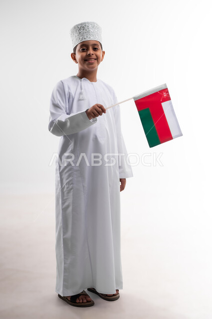 أعياد ومناسبات وطنية، إحياء ذكرى اليوم الوطني العماني، الاعتزاز والفخر بالرموز والراية العمانية، بورتريه لطفل عربي خليجي عماني يرتدي الدشداشة والكمه يحمل بيده علم صغير لسلطنة عمان، مفهوم حب الوطن والإنتماء إليه، الجسم بالطول الكامل، خلفية بيضاء