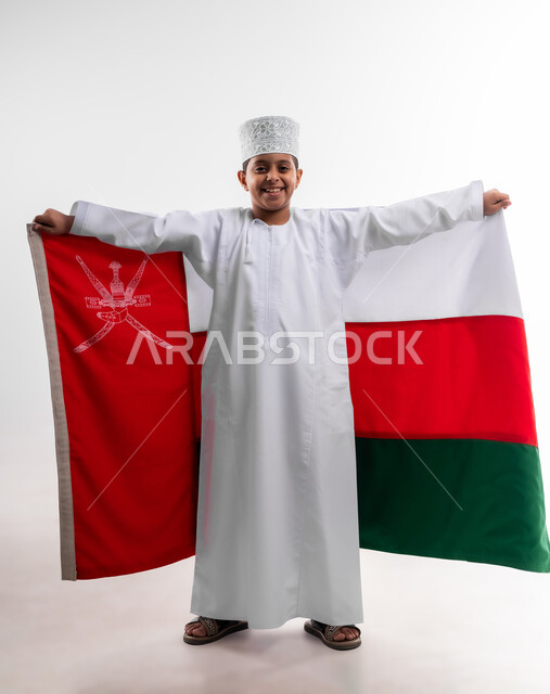 إحياء ذكرى اليوم الوطني العماني، الرايات والأعلام الوطنية للدول العربية، الاعتزاز والفخر بالرموز والراية العمانية، بورتريه لطفل عربي خليجي عماني يرتدي الدشداشة والكمه يضع علم سلطنة عمان على ظهره، مفهوم حب الوطن والإنتماء إليه، الجسم بالطول الكامل، خلفية بيضاء