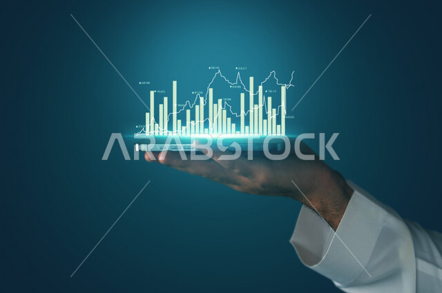 صورة مقربة لرجل عربي خليجي سعودي يحمل بيديه جهاز تقني ذكي عليه رسومات بيانية ثلاثية الأبعاد بتقنية الهولوجرام ،  صعود و نزول البورصة المالية ، الاستثمار المالي و الاقتصادي ، تطور و ابتكار .