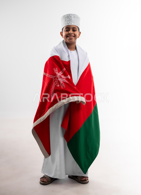 إحياء ذكرى اليوم الوطني العماني، الرايات والأعلام الوطنية للدول العربية، الاعتزاز والفخر بالرموز والراية العمانية، بورتريه لطفل عربي خليجي عماني يرتدي الدشداشة والكمه يضع علم سلطنة عمان على ظهره، مفهوم حب الوطن والإنتماء إليه، الجسم بالطول الكامل، خلفية بيضاء
