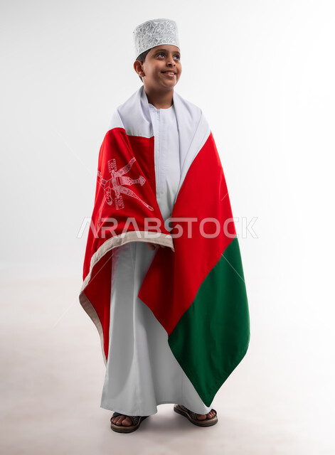 إحياء ذكرى اليوم الوطني العماني، الرايات والأعلام الوطنية للدول العربية، الاعتزاز والفخر بالرموز والراية العمانية، بورتريه لطفل عربي خليجي عماني يرتدي الدشداشة والكمه يضع علم سلطنة عمان على ظهره، مفهوم حب الوطن والإنتماء إليه، الجسم بالطول الكامل، خلفية بيضاء