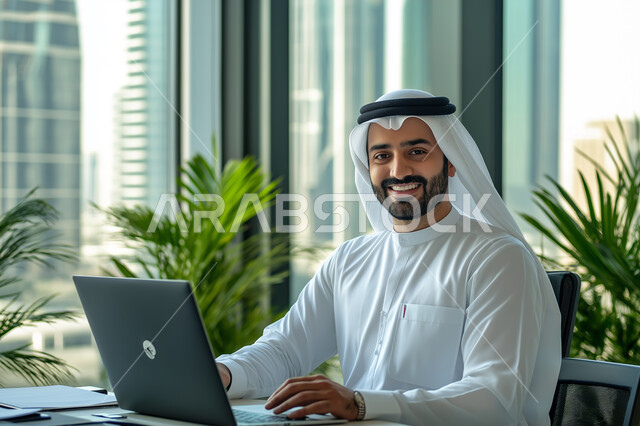 Business Administration and Organization, Image of a smiling Saudi Arabian Gulf man working on a computer inside the office, working to achieve goals and strategies, office administrative professions and jobs, using modern and advanced technological devices