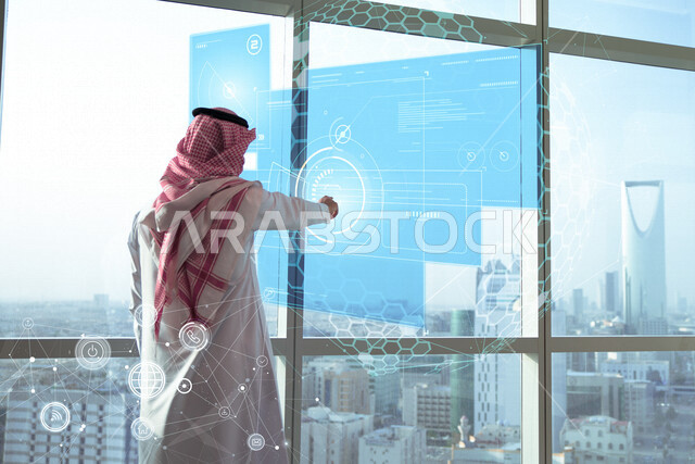 تصوير من الخلف لرجل اعمال سعودي خليجي يستخدم شبكة الكترونية بتقنية الهولجرام ، ايقونات خاصة بعملية الاتصال والتواصل الالكتروني ، شركة اعمال سعودية ، ابراج وناطحات سحاب