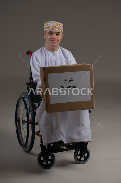 ذوي الإحتياجات الخاصة والهمم, بورتريه لشاب عربي خليجي عماني يرتدي الكمه والدشداشة ينظر الى الكاميرا بتعابير السرور ويحمل بيديه صندوق تبرع، مفهوم مساعدة المحتاجين، خلفية رمادي